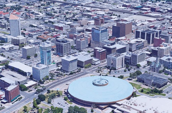 Aerial view of a cityscape featuring numerous mid-rise buildings, with a large round building with a blue roof in the foreground, surrounded by streets, parking lots, and trees.