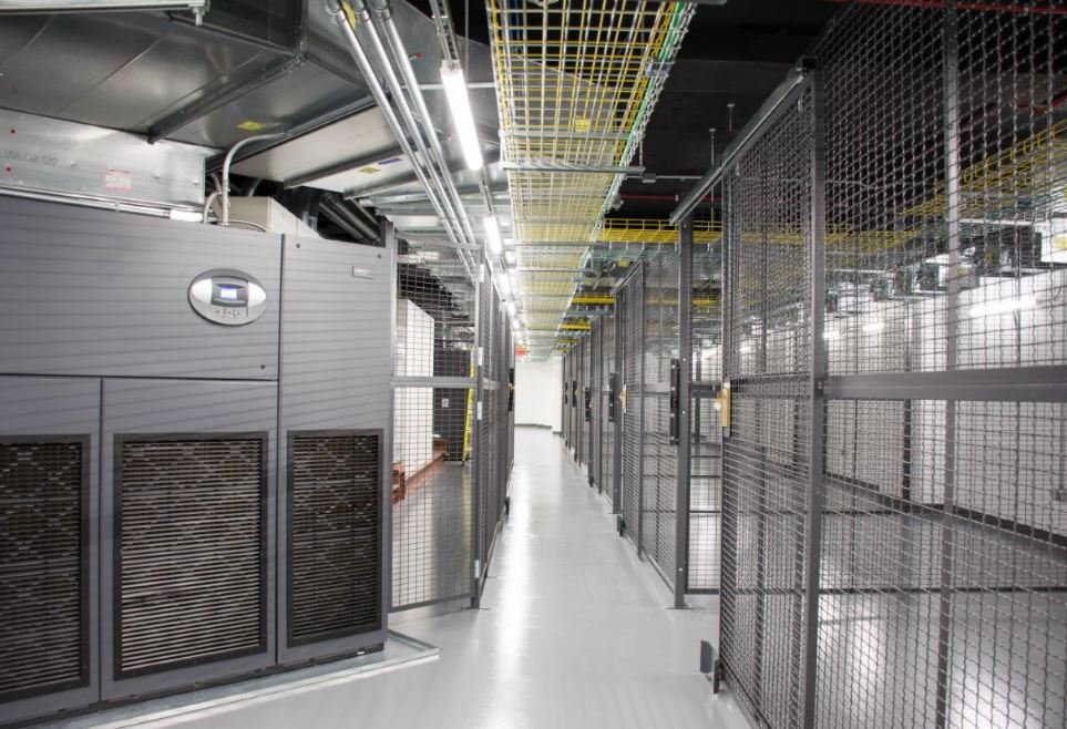 A secure data center corridor with metal mesh cages on the right, industrial cooling units on the left, overhead cable trays, and bright ceiling lights illuminating the white floor and walls.