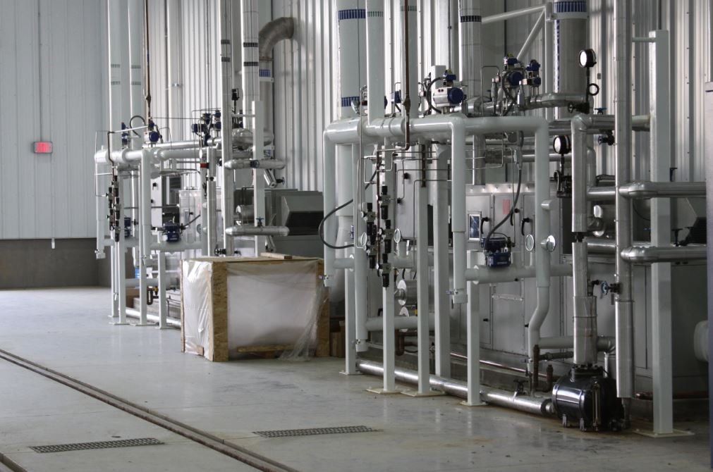 Industrial facility interior with metal pipes, valves, gauges, and machinery installed along the wall. A wooden crate with a plastic covering sits nearby. The floor is concrete, and the space is well-lit.
