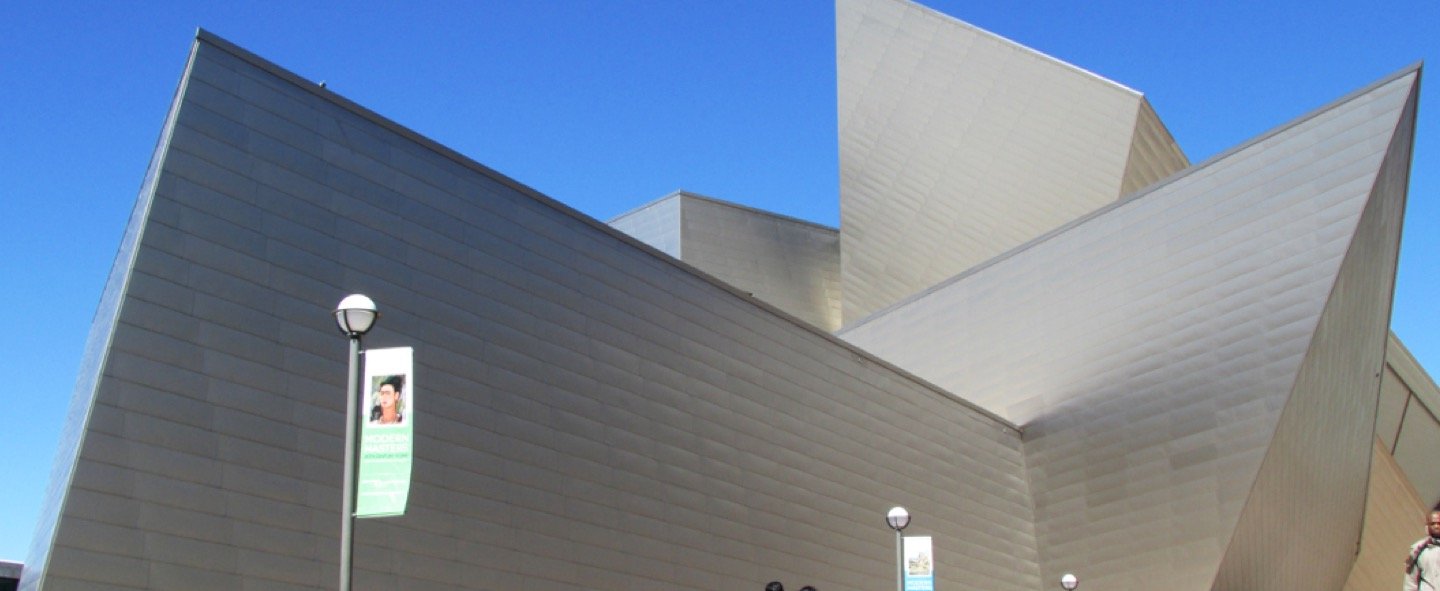 An angular, modern building with metallic, overlapping panels and sharp edges stands against a clear blue sky. Several round street lights and banners line the sidewalk in front.