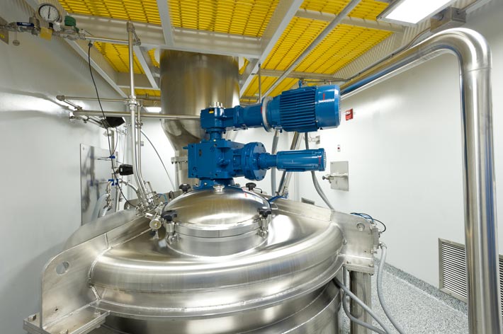 A large stainless steel industrial mixing tank with attached blue motorized equipment and surrounding pipes in a clean, modern facility with yellow ceiling grates.