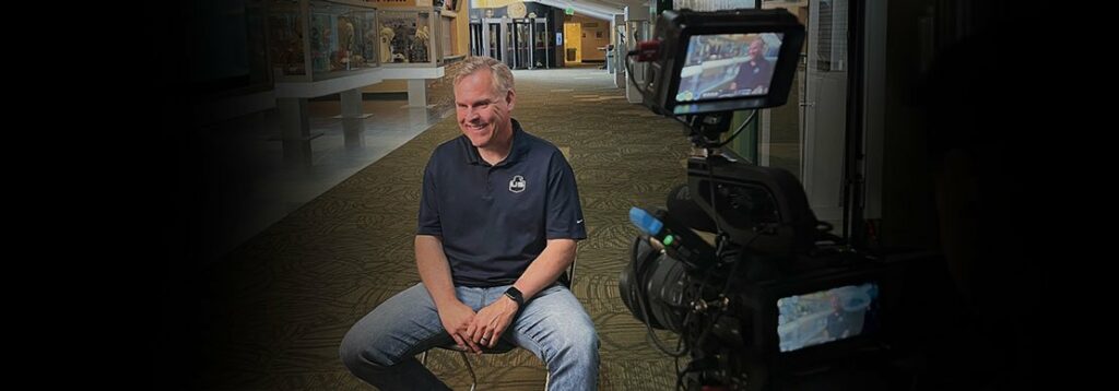 A man in a dark polo shirt and jeans sits on a stool, smiling, while being filmed by a video camera in a hallway with display cases and overhead lights.
