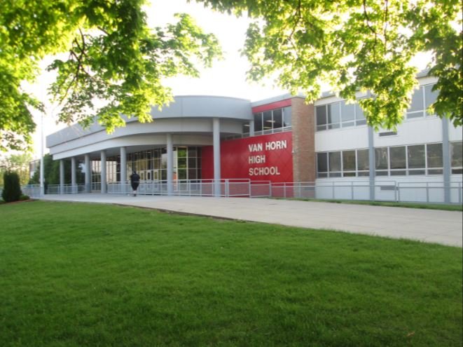 A modern school building with large windows and a red sign reading Van Horn High School, set behind a green lawn and shaded by leafy trees on a sunny day.