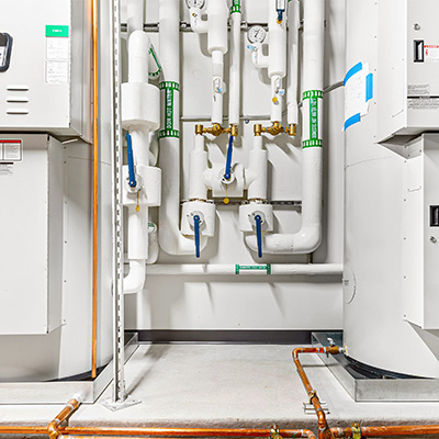 A mechanical room with white insulated pipes, blue-handled valves, gauges, and labeled sections. Copper pipes run along the floor, and large equipment units are visible on both sides of the image.