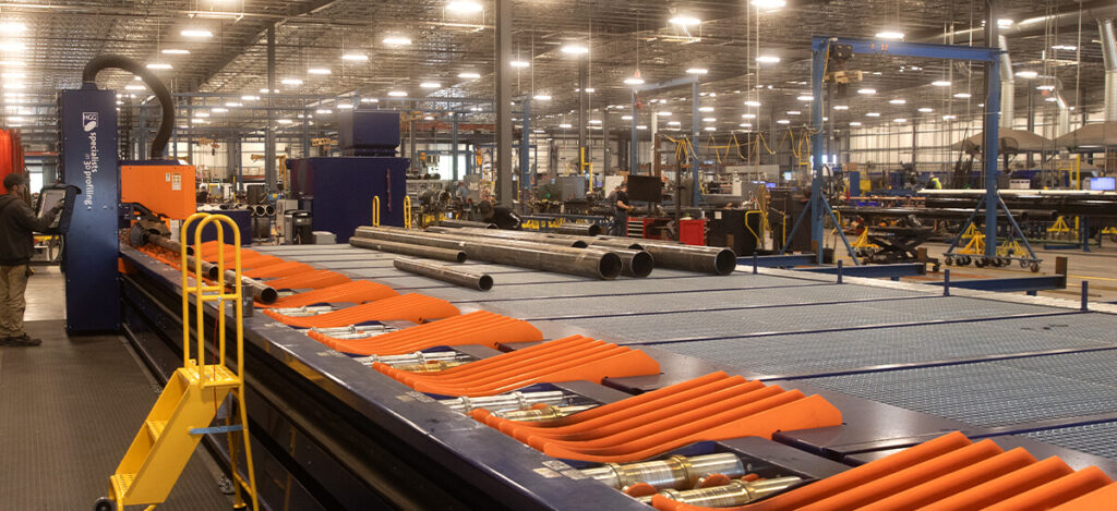 A large industrial facility with metal pipes, machinery, and orange equipment. Overhead lights illuminate the spacious interior, where various machines and tools are arranged for manufacturing or fabrication work.